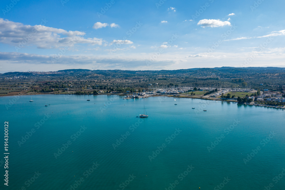 Obraz premium Aerial view of a peaceful marina and turquoise bay near Kilada, Greece. Calm water reflects scattered sailboats, with rolling hills and a partly cloudy sky in the background.