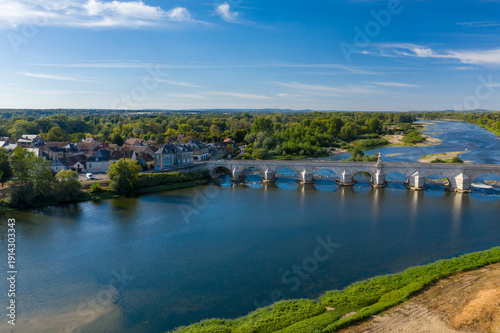 Wide aerial perspective of La Charite sur Loire in central France, featuring a historic stone bridge crossing the Loire river, traditional rooftops, and lush greenery along the riverside. The scene