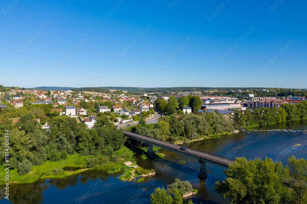 Fototapeta premium High-angle panorama of Imphy in central France, showing a bridge crossing the river, lush green trees, and residential rooftops under a vivid blue sky. The image highlights the tranquil rural
