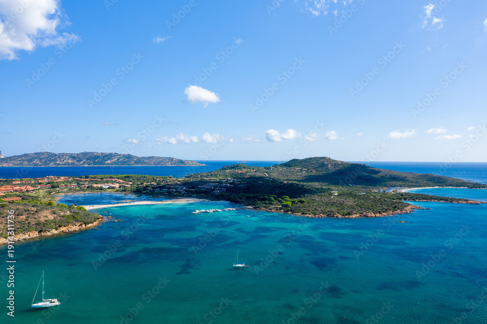 Fototapeta premium Drone perspective over Spiaggia Salina Bamba near San Teodoro, Sardinia, revealing vivid turquoise bays, anchored sailboats, and lush green hills under a clear blue sky.