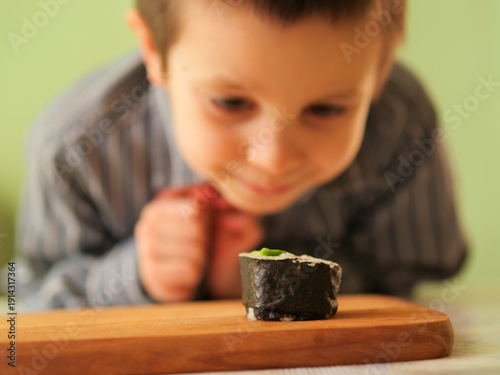 young boy expressing joy and curiosity while examining a sushi roll in a home kitchen - single handmade maki roll on a wooden board