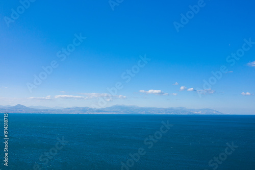 Fotografie Wide open seascape featuring deep blue water and a faint mountain range on the horizon, all beneath a cloud-dotted, vibrant sky