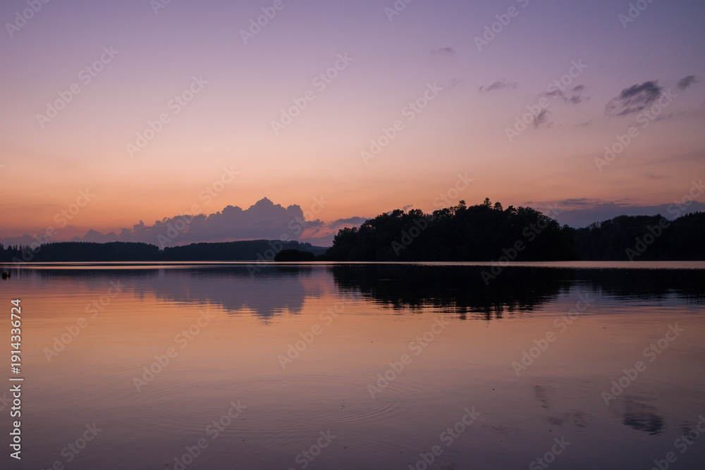 Fototapeta premium Soft morning light illuminates calm waters at Lac des Settons, with gentle pastel colors in the sky and a dark forest silhouette mirrored on the tranquil lake surface.