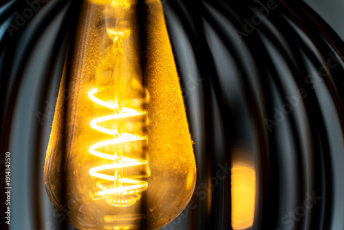 Spiral warm filament of a glowing bright light bulb inside a lampshade. The illuminated coil creates a cozy and inviting atmosphere with soft amber light.