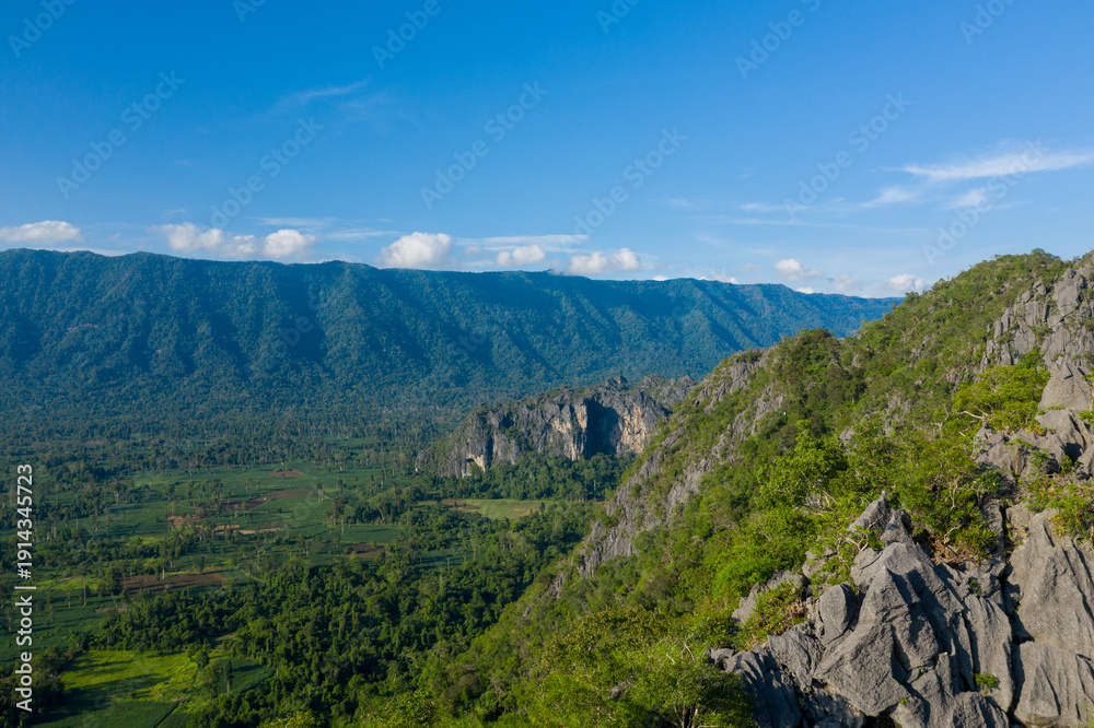 Fototapeta premium Expansive view of a rugged limestone ridge covered in lush vegetation, overlooking a wide green valley and distant forested mountains under a clear blue sky. The scene features sharp rock textures