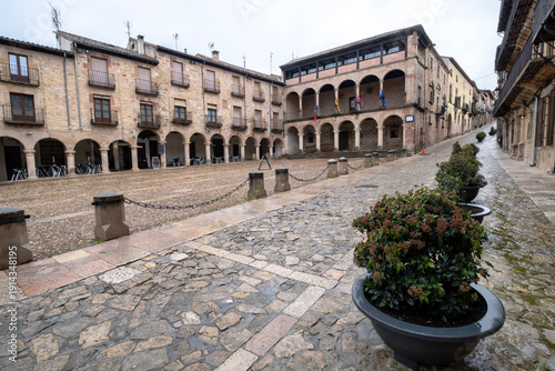 Wide angle view of the historic Plaza Mayor in Siguenza Spain featuring traditional stone buildings with arched galleries and empty cobblestone pavement under a cloudy winter sky in castile