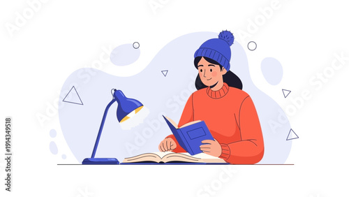 Young woman in winter hat reading book under desk lamp, studying at home