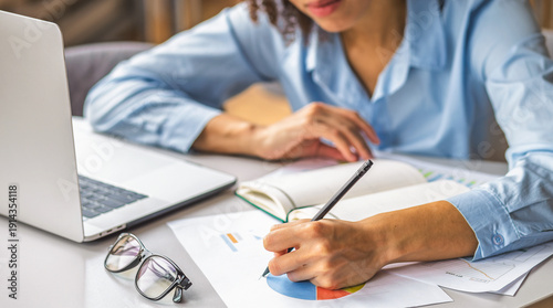 Woman analyzing financial report and writing notes on paper charts beside laptop. Business planning, data analysis, strategy, accounting, productivity, home office