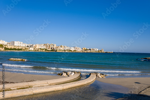 Canvas Print Modern white buildings line the Otranto waterfront along the turquoise Adriatic Sea, with gentle waves lapping a sandy beach in bright daylight