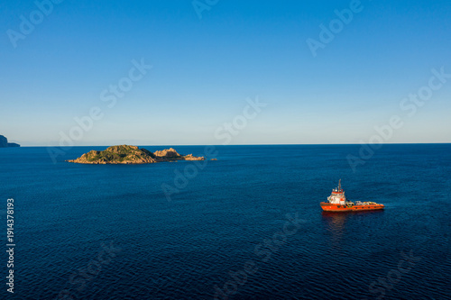 Canvas Print A vibrant red workboat floats on deep blue Mediterranean water near the rocky green Isola dell'Ogliastra, with clear skies and distant coastline visible near Lotzorai, Sardinia