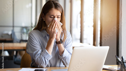 Young woman at an office desk sneezes into a tissue while working on a laptop, illustrating cold or allergy symptoms, workplace illness, discomfort and the need for hygiene, rest and self-care.

