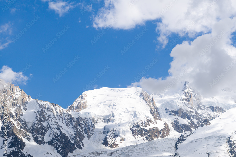 Fototapeta premium Sunlight bathes the pristine, snow-covered crest of Mont Blanc du Tacul, with rugged rock faces and drifting clouds set against a vivid blue alpine sky.