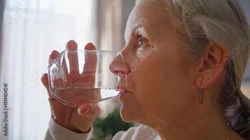 Senior woman takes painkiller with water sip in kitchen closeup