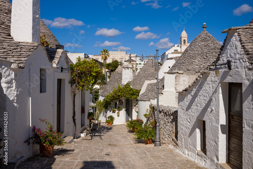 Traditional whitewashed trullo houses with conical stone roofs line a narrow cobblestone alley in Alberobello, Apulia, Italy. Potted plants, rustic textures, and clear blue sky evoke the charm of