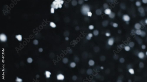 Beautiful macro close-up footage captures soft white snow falling gently against a deep, dark nighttime background creating abstract bokeh light effects and winter atmosphere.