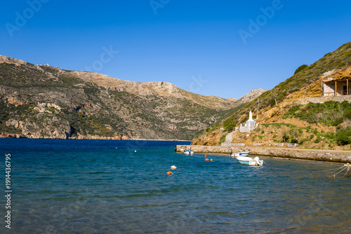 Small boats are moored along a stone pier in the sheltered blue bay of Porto Kagio, with rugged hills and sunlit rocky slopes rising in the background under a clear sky.