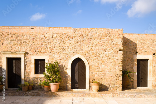 Sunlit stone building with an arched entrance and potted plants, featuring textured walls and simple geometric shapes under a clear blue sky in Crete. The scene feels warm, inviting, and authentically