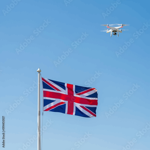 Union Jack flag of the United Kingdom waving proudly on a flagpole with a modern drone flying in the clear blue sky with white clouds.