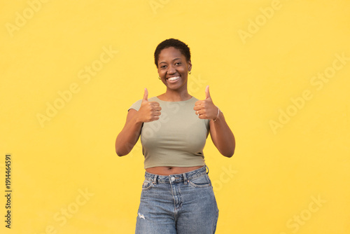 Portrait of young woman with short Afro hair showing thumbs up with both hands on yellow background. Positive emotion and cheerful mood