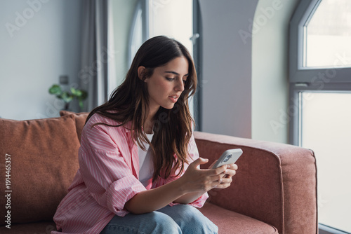 Quadro su tela A confused and angry young woman sits on the couch at home looking at her smartp