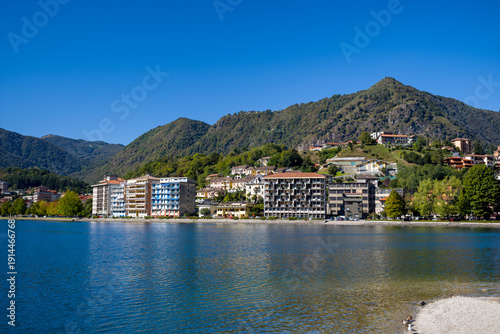 Tableau sur toile Modern lakeside buildings and hillside houses line the shore of Omegna on Lake Orta in northern Italy, with forested mountains rising in the background under a vivid blue sky