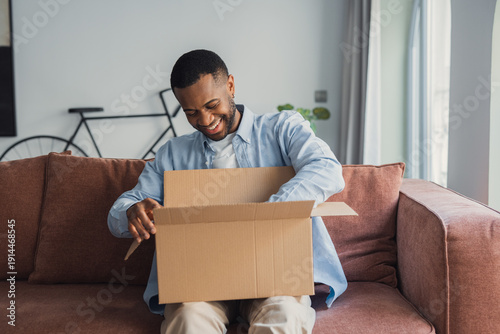 Bilde på lerret Smiling African American cheerful man unpacking parcel, sitting on couch at home