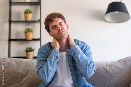 Valokuva An upset and tired young man sits at home massaging his stiff neck and tense sho