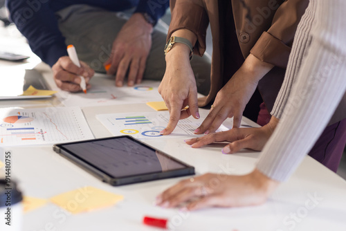 Diverse coworkers collaborating over printed charts and tablet computer around office meeting table