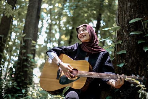 Happy young woman playing acoustic guitar outdoors amidst lush green trees and sunlight