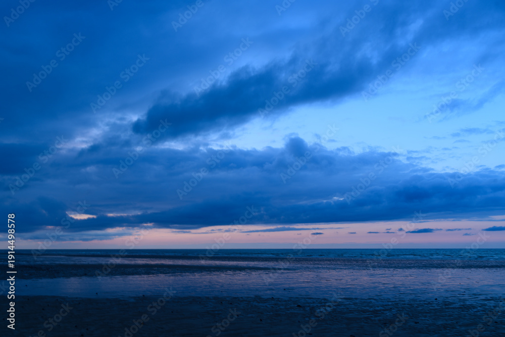 Obraz premium Moody twilight clouds stretch across the sky above the calm, reflective waters of the English Channel, with soft blue and pink hues creating a tranquil seascape at low tide.