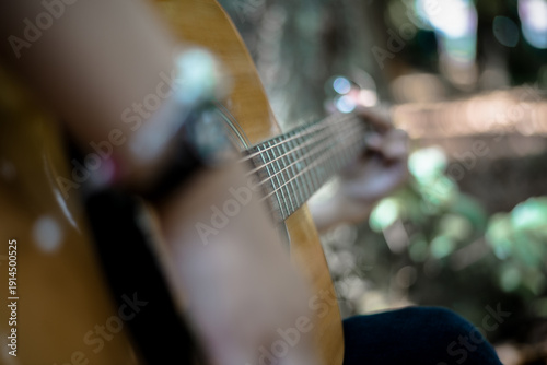 Person playing acoustic guitar outdoors among blurred green foliage and warm sunlight