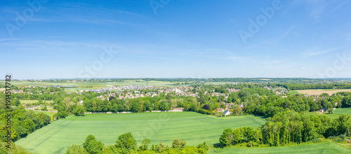 Wide drone panorama capturing lush green fields, dense tree lines, and the village of Ranville under a vivid blue sky with expansive rural views.