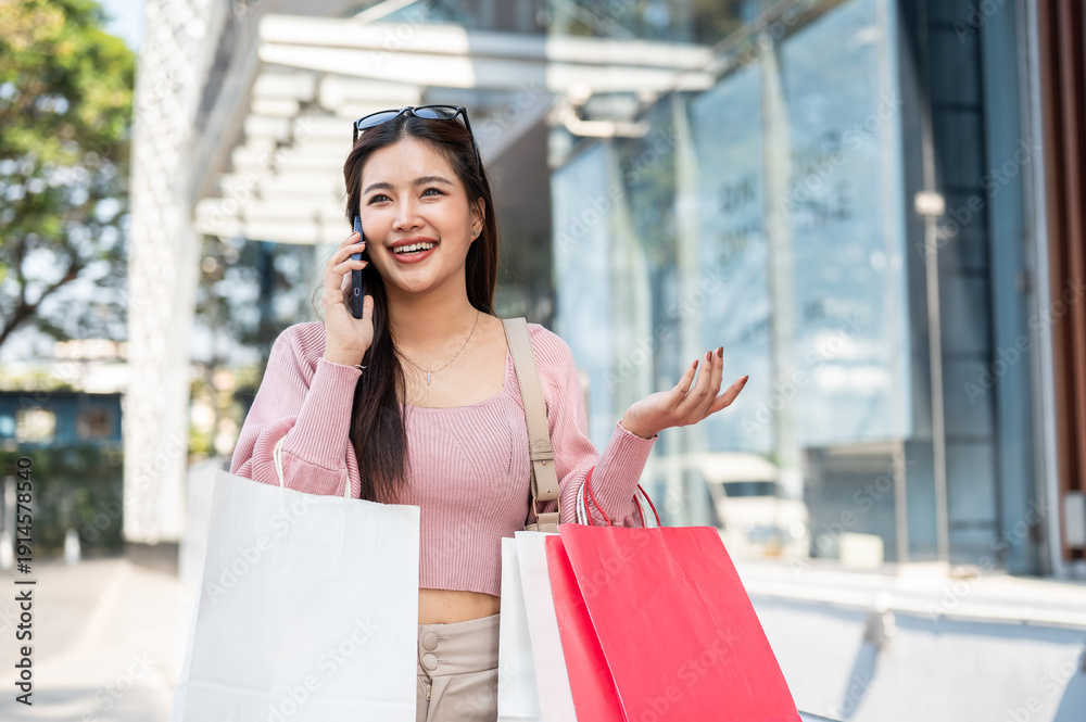 Fototapeta premium Pretty asian woman carrying shopping bag talking&nbsp;on&nbsp;phone&nbsp;while standing walking outside a mall.