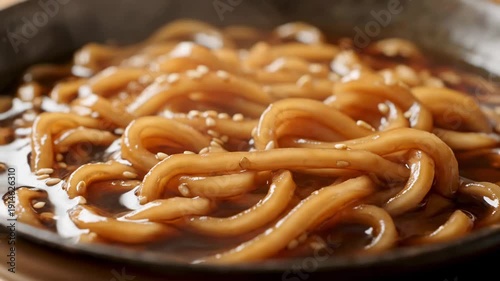 Close-up of thick, glossy Asian noodles coated in a savory brown sauce and sesame seeds being lifted or stirred in a dark bowl, emphasizing the appetizing texture for a culinary advertisement.