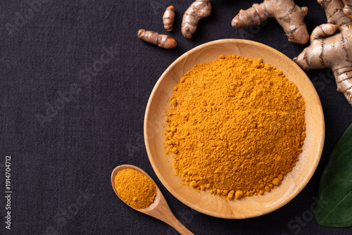 Top view of turmeric powder in a wooden bowl with a wooden spoon and fresh turmeric roots on black fabric background. Organic healthy spice