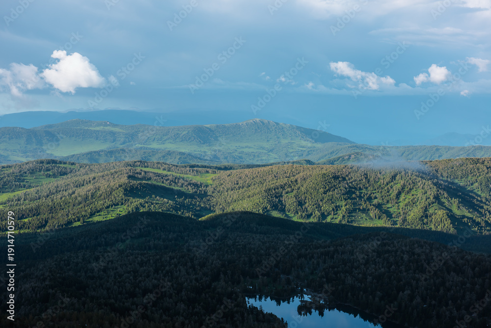 Fototapeta premium Scenic aerial top view above forest lake to green hilly vastness and layered mountain silhouettes far away under low dramatic cloudy sky. Wonderful scenery with sunlight and shadows of clouds on hills