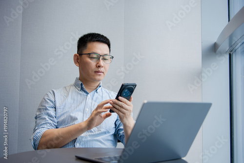 Photography A bespectacled man in an office uses his smartphone while looking at a laptop co