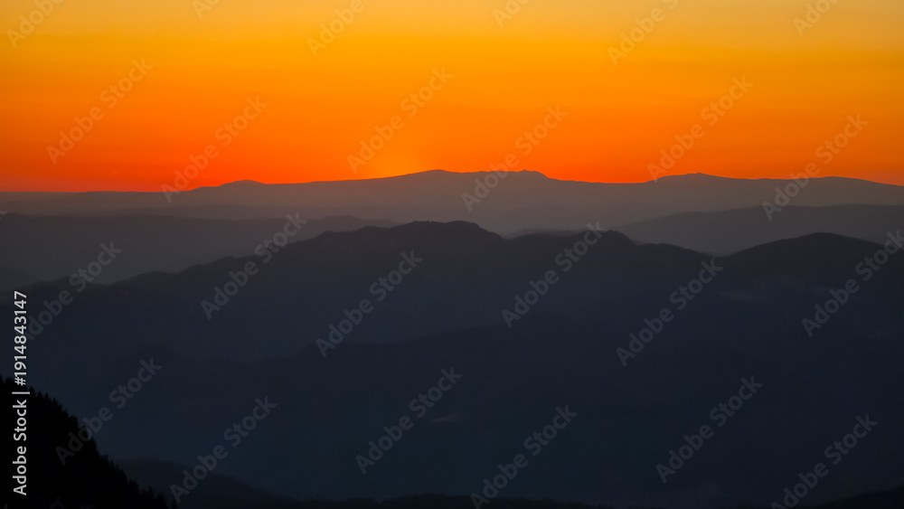 Obraz premium Rolling silhouettes of the Carpathian Mountains at sunset, with hazy layers of ridges fading into the vibrant, golden-orange sky. A tranquil, majestic nature background.