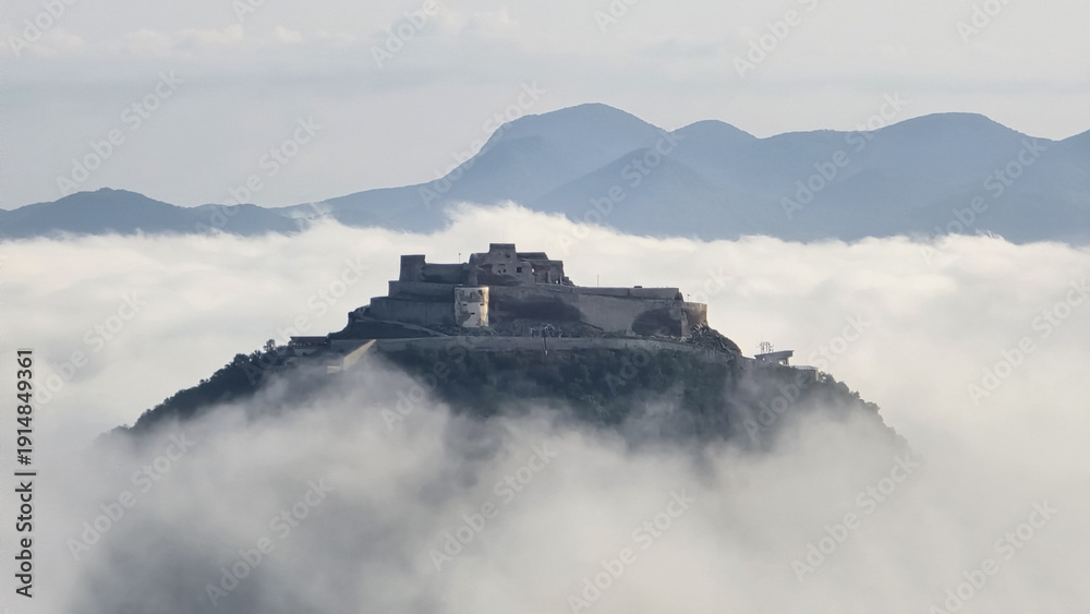 Fototapeta premium The Fortress of Deva, Romania, sits atop a hill, partially shrouded in a sea of morning fog, with distant Carpathian mountains visible in the hazy background under a soft, diffused light.