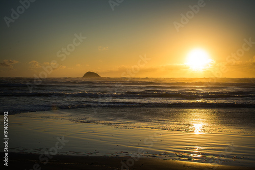 Golden hour descends over Muriwai Beach, casting warm light across gentle waves and reflective sands, while the iconic offshore rock rests silhouetted beneath a sky of drifting clouds and seabirds