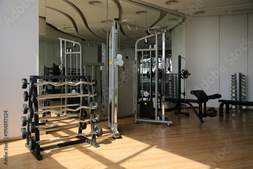 Interior view of modern fitness center gym with equipment. Home sport training room featuring weight rack barbells and cable machine with mirror wall and sunlight on wooden floor background.