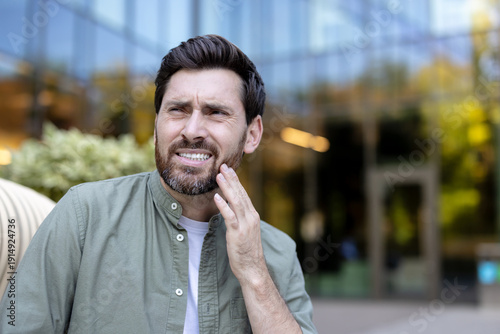 Fotografie Man grimacing outdoors, holding cheek in pain from a toothache, showing dental d
