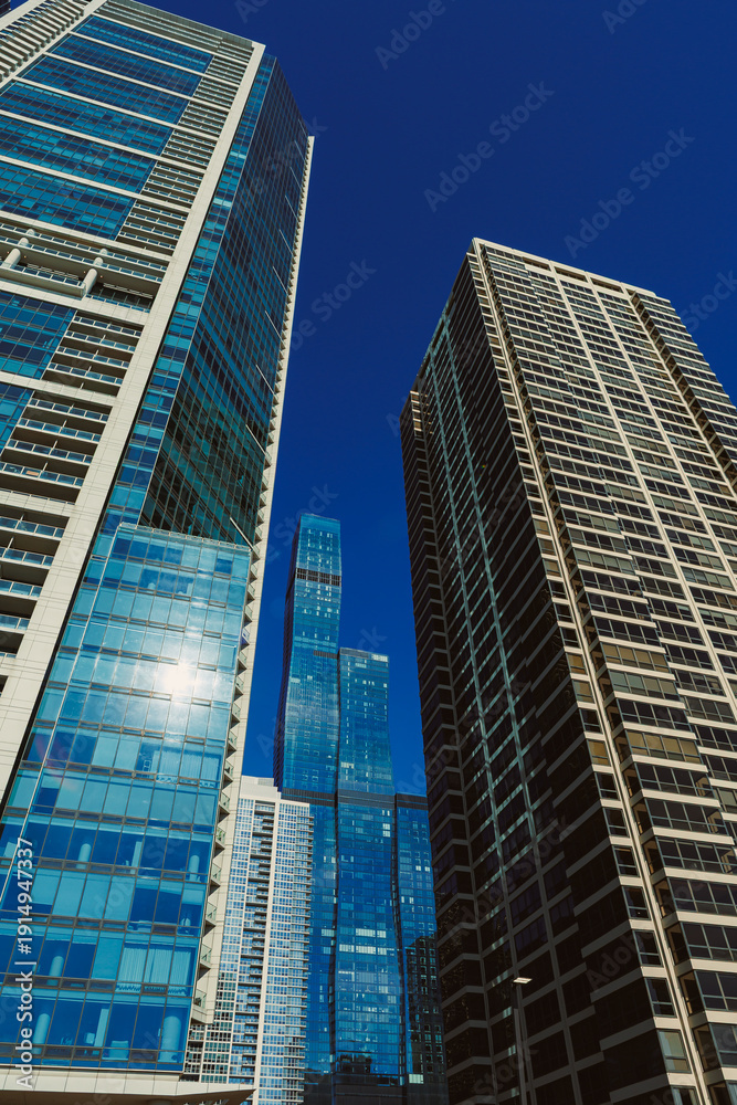 Fototapeta premium Tall buildings reach upward in a city skyline during midday with a clear blue sky and sunlight reflecting off glass windows