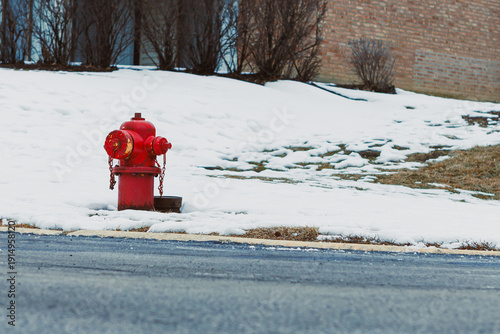 Bright red fire hydrant stands beside snowy ground in winter season near a residential area with grass patches showing
