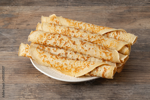 Stack of thin crepes on a white plate placed on a wooden table next to a stone wall in a kitchen setting during daylight hours