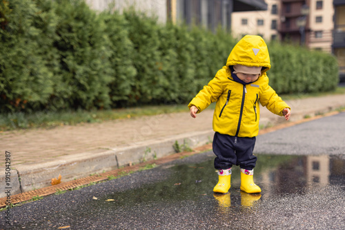 Happy toddler walking in puddles, little boy in bright clothes enjoying walk on rainy day