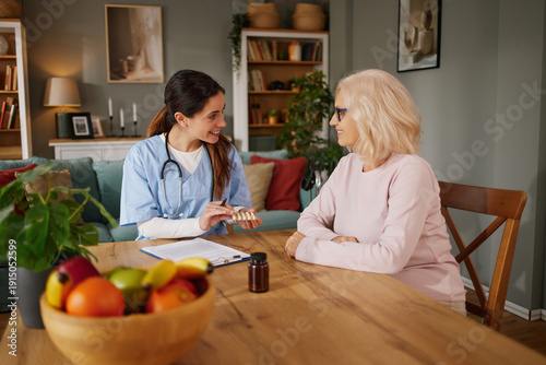 A home health nurse talks to a senior patient about medications while sitting at a table. The setting features a homey atmosphere with plants and a comfortable couch in the background.