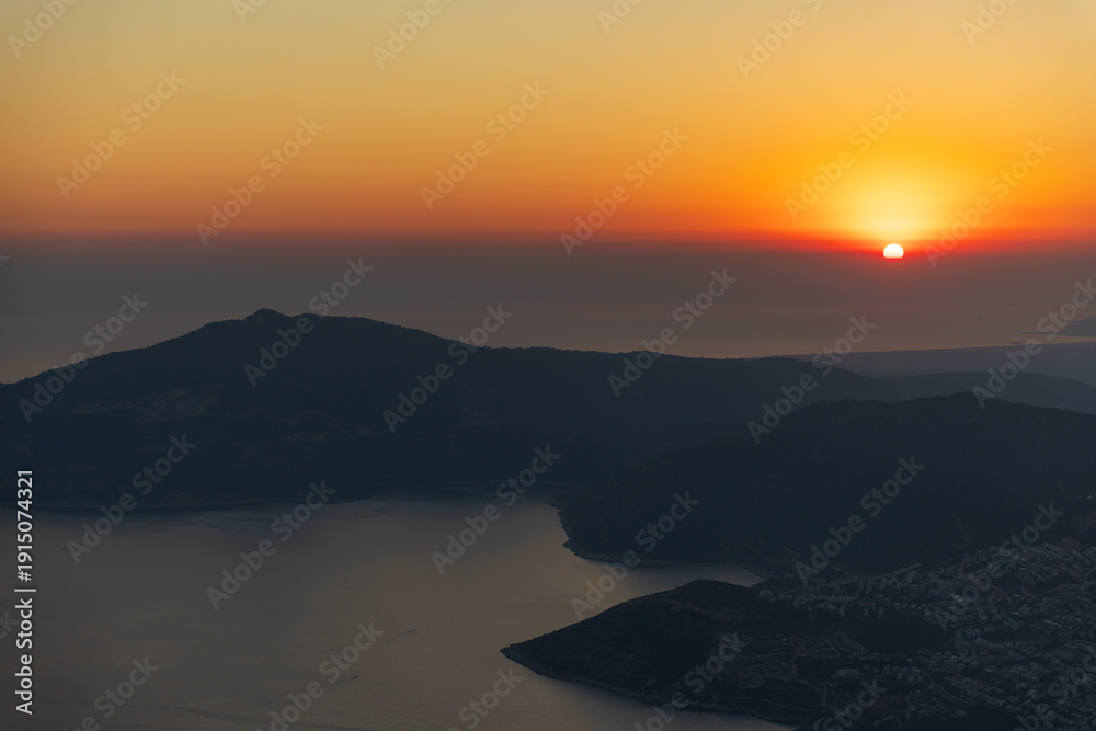 Fototapeta premium Tranquil sunset slides below dark distant mountains, casting warm evening light over a coastal valley and calm sea. Silhouettes and mist create a peaceful, scenic landscape. Turkey