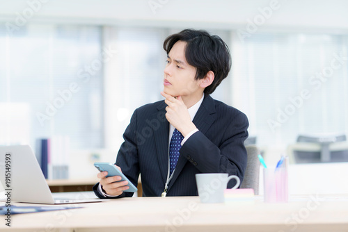 オフィスでノートパソコンで仕事をする困った顔のスーツを着た若い日本人男性