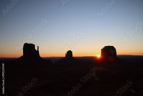 Mitten Buttes and Merrick Butte in Monument Valley, USA, at sunrise with the sun visible on the horizon. The sandstone formations appear as dark silhouettes against a warm orange sky.
